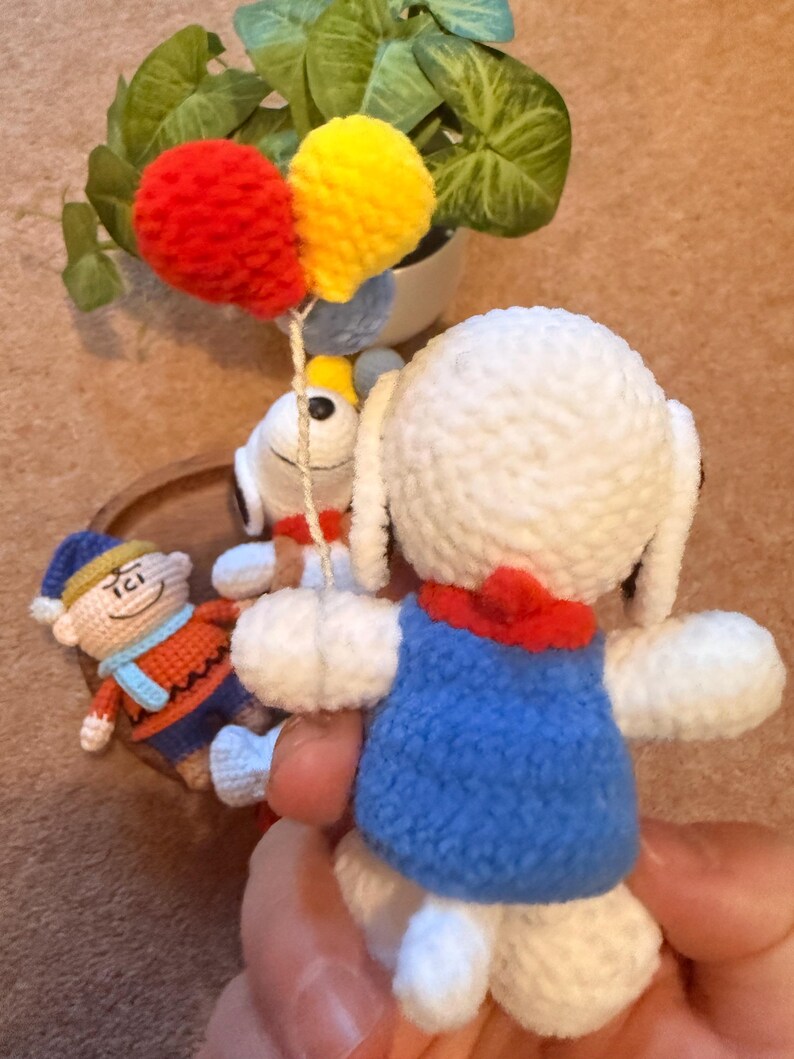 May include: Handmade crochet Snoopy doll wearing a blue vest and red collar. The doll is white with black eyes and ears. It is holding a string with red and yellow crochet balloons. A small crochet figure of Charlie Brown is in the background.