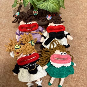 May include: Four handmade crochet dolls with exaggerated red lips, each with unique outfits and hairstyles. The dolls have brown yarn hair, and wear dresses in white, green, and purple. One doll has a black purse. The dolls are arranged in front of a green plant.