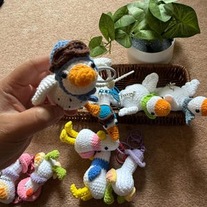 Puede incluir: Una colecci&oacute;n de llaveros de patos de ganchillo hechos a mano en varios colores. Un pato se sostiene en una mano, con un sombrero de aviador azul y marr&oacute;n. Otros patos est&aacute;n en una cesta, y algunos est&aacute;n tumbados. Los patos tienen picos naranjas y acentos coloridos.