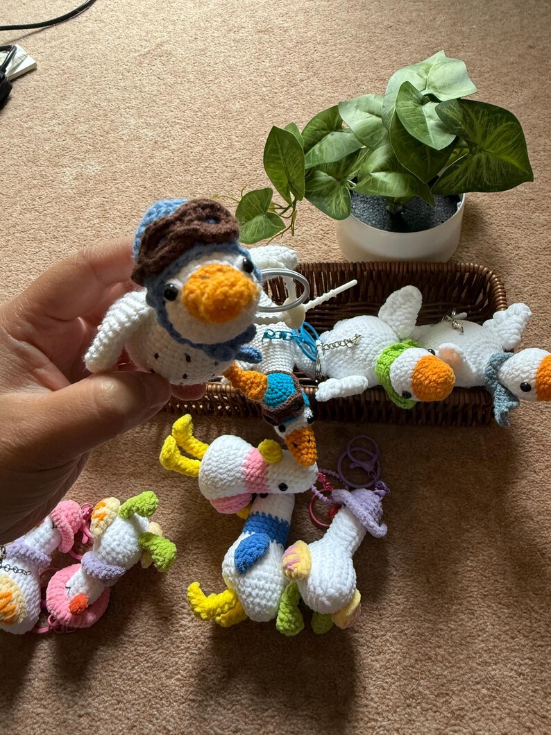 Puede incluir: Una colecci&oacute;n de llaveros de patos de ganchillo hechos a mano en varios colores. Un pato se sostiene en una mano, con un sombrero de aviador azul y marr&oacute;n. Otros patos est&aacute;n en una cesta, y algunos est&aacute;n tumbados. Los patos tienen picos naranjas y acentos coloridos.
