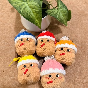 May include: Five handmade crochet keychains shaped like whimsical characters, each with a different coloured hat: blue, red, orange, yellow, and pink. The keychains have black eyes, red accents, and a small loop for attaching keys.