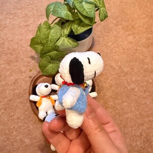 May include: Handmade crochet Snoopy figurines. The main figurine is white with black ears, a blue vest, and a red collar. A small bell hangs from the collar. Other Snoopy figurines are in a wooden heart-shaped bowl. A green plant in a white pot is in the background.