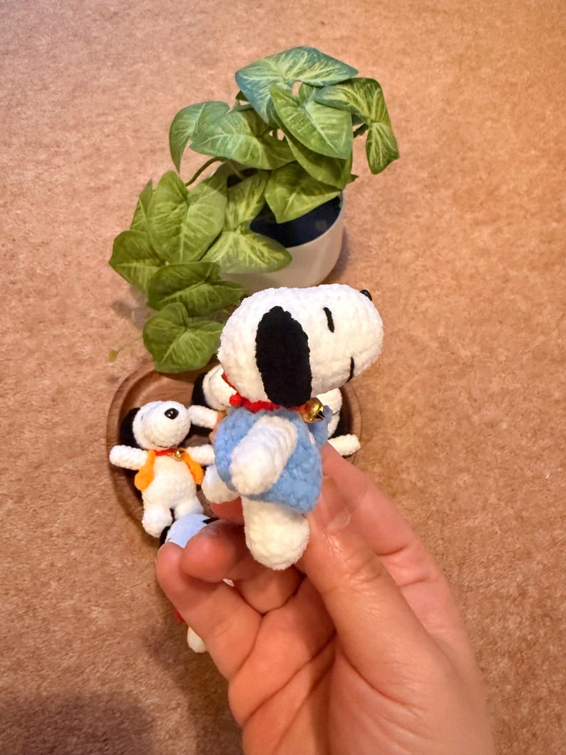 May include: Handmade crochet Snoopy figurines. The main figurine is white with black ears, a blue vest, and a red collar. A small bell hangs from the collar. Other Snoopy figurines are in a wooden heart-shaped bowl. A green plant in a white pot is in the background.