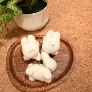 May include: Four small, white felt bunny figures with simple embroidered faces are arranged on a wooden plate. A green ribbon is tied around one bunny. A white potted plant with green leaves is in the background.