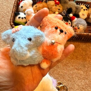 May include: A light blue elephant and orange fox plush keychains are held in a hand, with a basket of other plush animals in the background. The elephant has a pink flower and the fox has a small bell. The keychains are made of soft, fluffy material.