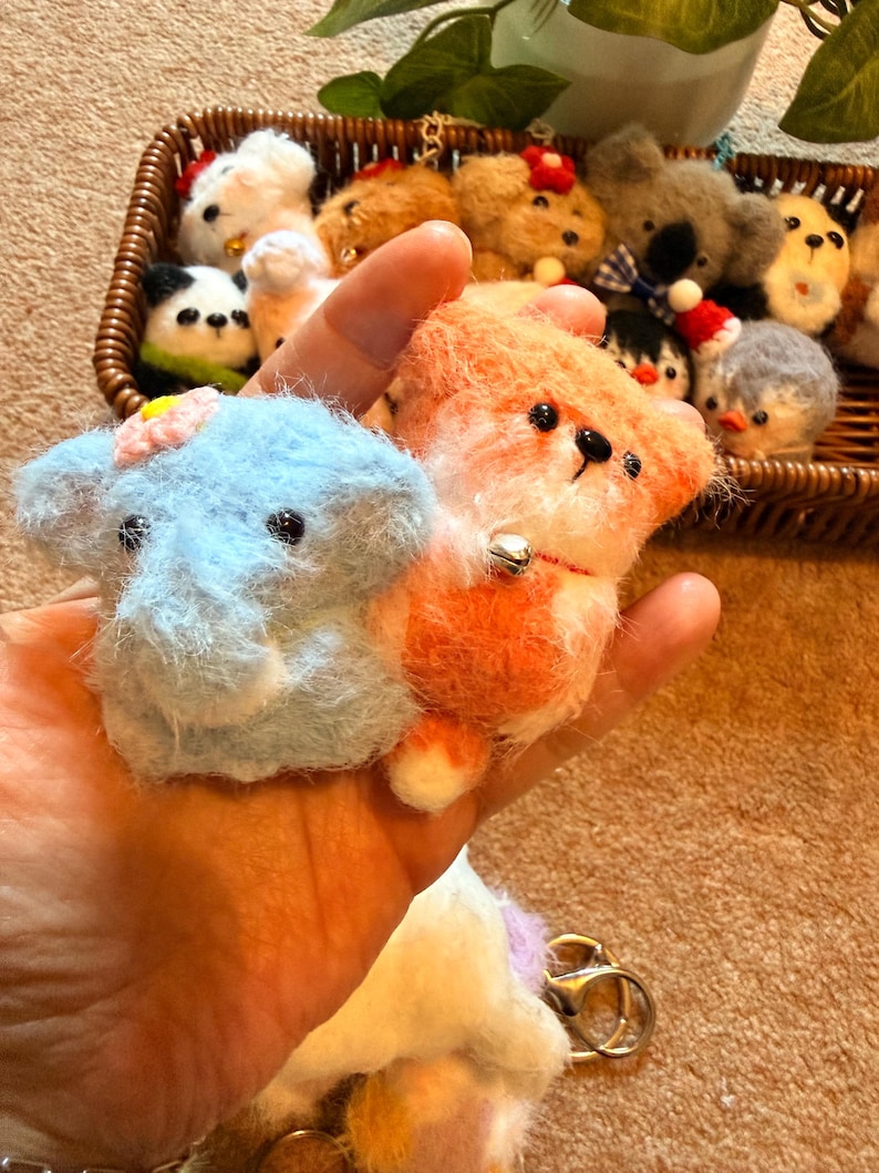May include: A light blue elephant and orange fox plush keychains are held in a hand, with a basket of other plush animals in the background. The elephant has a pink flower and the fox has a small bell. The keychains are made of soft, fluffy material.