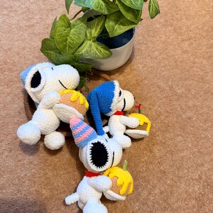 May include: Crocheted Snoopy figurines, each holding a small cake. One Snoopy wears a party hat. The figures are white with black eyes and ears, and red collars. A potted plant with green leaves is in the background.