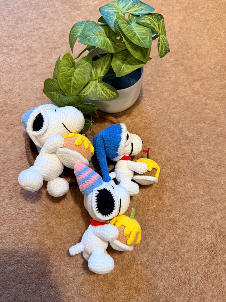 May include: Crocheted Snoopy figurines, each holding a small cake. One Snoopy wears a party hat. The figures are white with black eyes and ears, and red collars. A potted plant with green leaves is in the background.