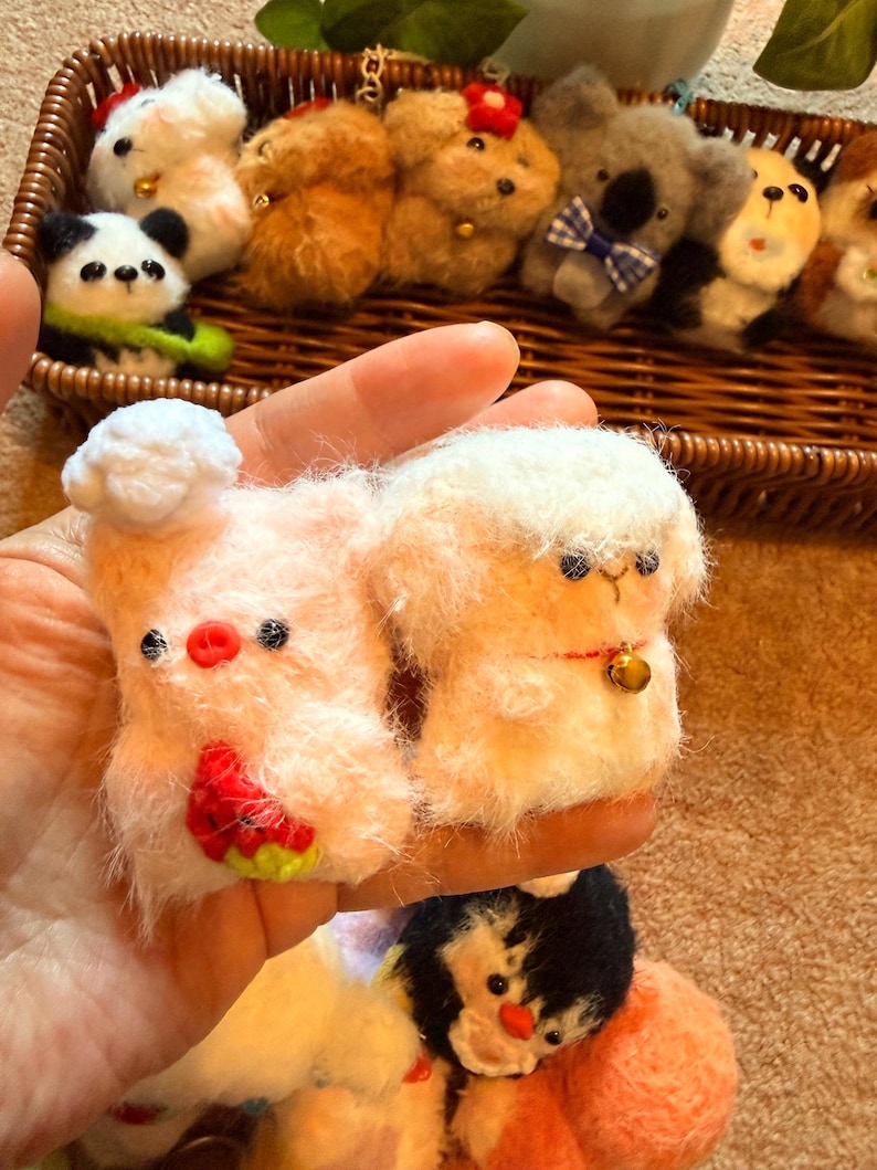 May include: A collection of handmade felt animal keychains, including a panda, koala, and various other creatures. The keychains are in a woven basket. The keychains are in various colors, including white, pink, brown, and black. The pig keychain has a watermelon design.