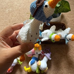 Puede incluir: Llaveros de ganso de ganchillo hechos a mano en varios colores. Un ganso lleva un sombrero y una bufanda de aviador azules. Otros gansos tienen detalles coloridos como picos naranjas, patas amarillas y detalles rosas, azules y morados. Los llaveros son blancos.