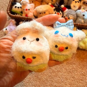 May include: A collection of handmade plush animal keychains. The keychains include a white sheep with a chick, and a white character with a blue bow. Other keychains in a basket include a panda, dogs, and other animals.