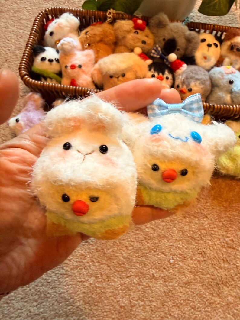 May include: A collection of handmade plush animal keychains. The keychains include a white sheep with a chick, and a white character with a blue bow. Other keychains in a basket include a panda, dogs, and other animals.