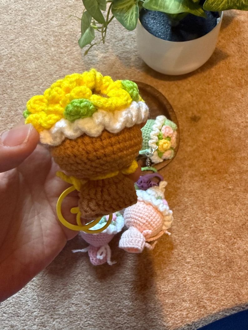 Pu&ograve; includere: Portachiavi a forma di cupcake all'uncinetto con glassa gialla, verde e bianca. Il cupcake ha una base marrone e un portachiavi giallo. Sono visibili anche altri cupcake all'uncinetto rosa e verdi.