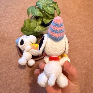 May include: Two handmade crochet Snoopy figures. One figure is lying down and holding a piece of cake. The other figure is upright and wearing a blue and pink striped party hat. A small green plant is in the background.