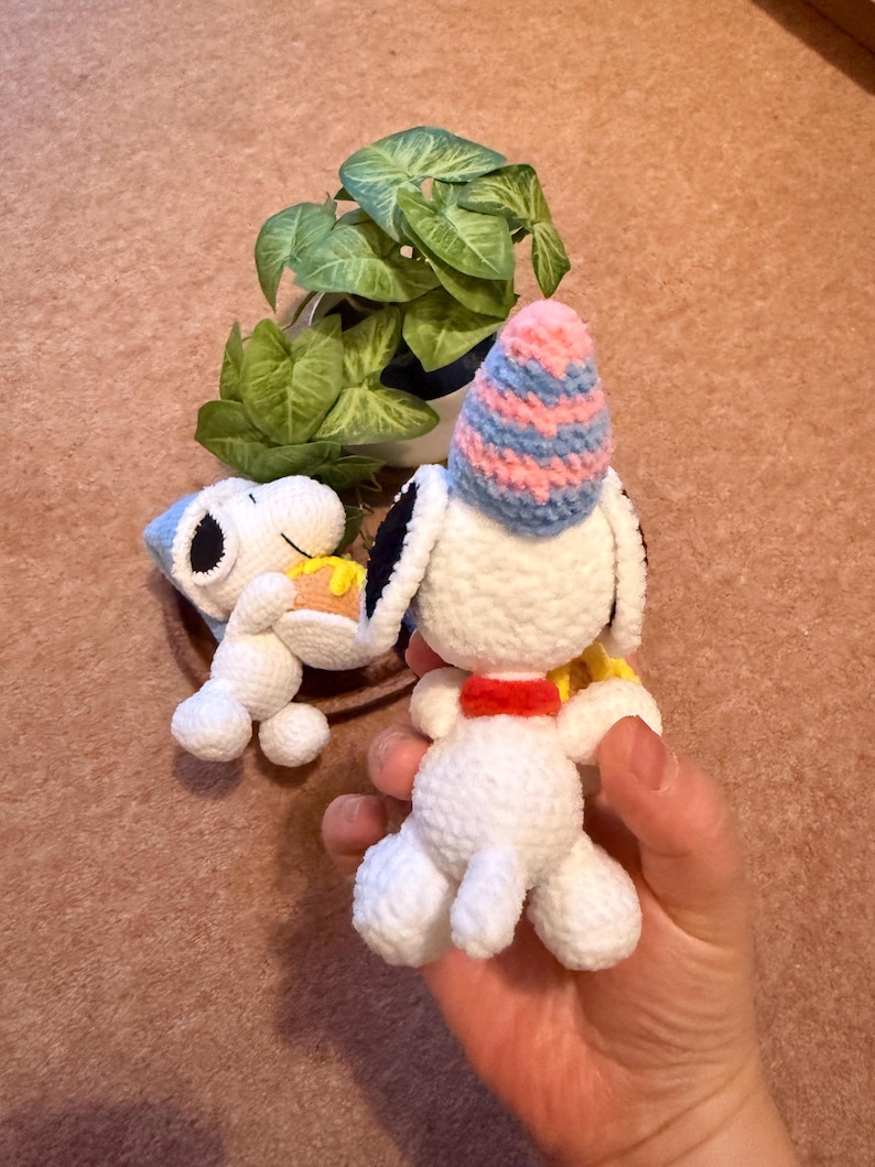 May include: Two handmade crochet Snoopy figures. One figure is lying down and holding a piece of cake. The other figure is upright and wearing a blue and pink striped party hat. A small green plant is in the background.