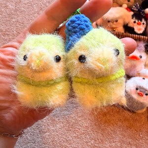 May include: Two handmade, fuzzy, yellow-green bird-shaped keychains with black eyes and green yarn accents. A blue crocheted loop is attached to the top of each bird. Other similar plush toys are visible in the background.