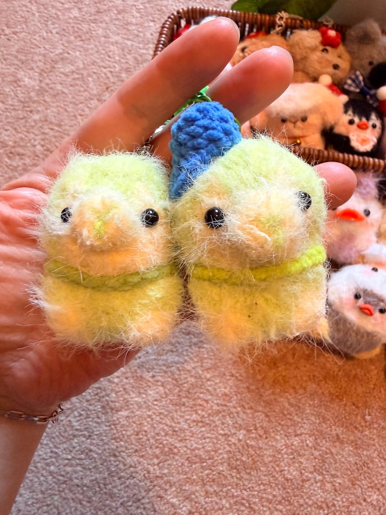 May include: Two handmade, fuzzy, yellow-green bird-shaped keychains with black eyes and green yarn accents. A blue crocheted loop is attached to the top of each bird. Other similar plush toys are visible in the background.