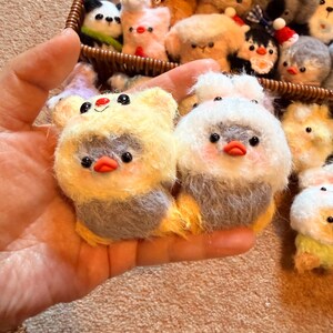 May include: A collection of handmade felt animal ornaments. The foreground features two plush bird-like figures with yellow and white fur, gray bodies, and orange beaks. A wicker basket in the background holds various other animal ornaments.