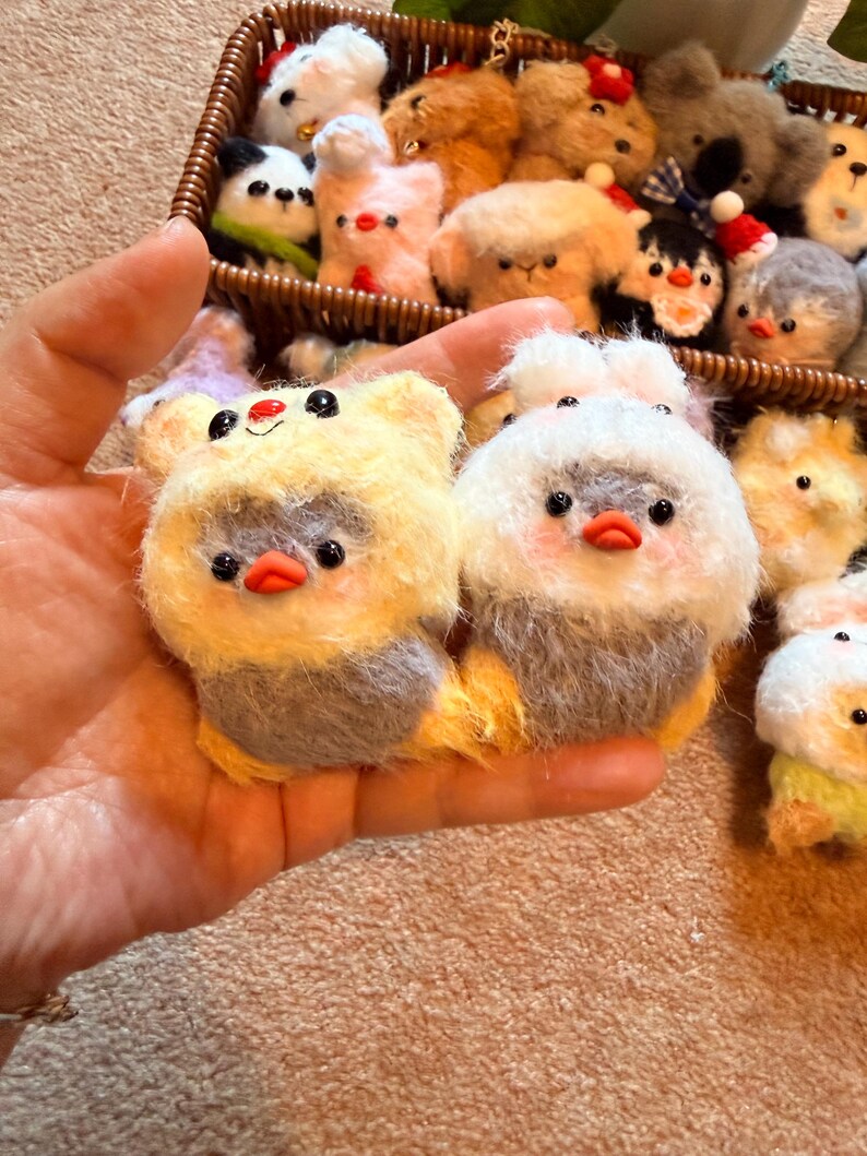 May include: A collection of handmade felt animal ornaments. The foreground features two plush bird-like figures with yellow and white fur, gray bodies, and orange beaks. A wicker basket in the background holds various other animal ornaments.