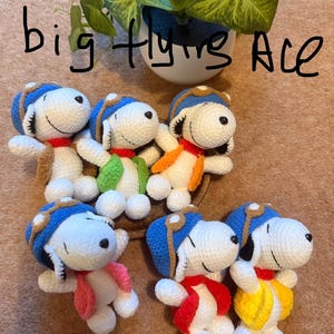 May include: Handmade crochet Snoopy dolls, each wearing a blue aviator cap and colorful scarves and vests. The text "big flying Ace" is written in black marker above the dolls. A green plant in a white pot is in the background.