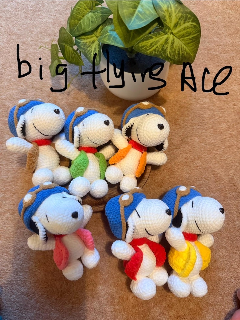 May include: Handmade crochet Snoopy dolls, each wearing a blue aviator cap and colorful scarves and vests. The text "big flying Ace" is written in black marker above the dolls. A green plant in a white pot is in the background.
