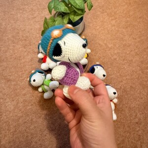 May include: Handmade crochet Snoopy dolls in various sizes. The dolls feature a white body, black ears, and a blue aviator helmet with orange goggles. One doll is held in a hand, showcasing the purple vest and white legs. A small green plant is in the background.