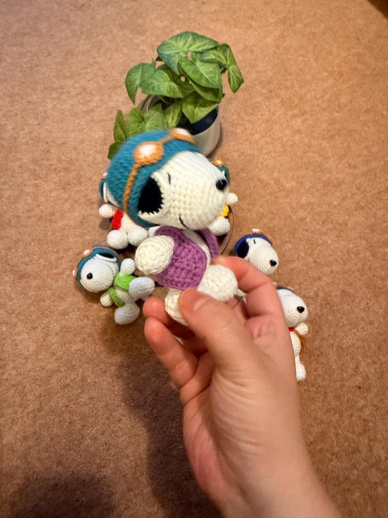 May include: Handmade crochet Snoopy dolls in various sizes. The dolls feature a white body, black ears, and a blue aviator helmet with orange goggles. One doll is held in a hand, showcasing the purple vest and white legs. A small green plant is in the background.
