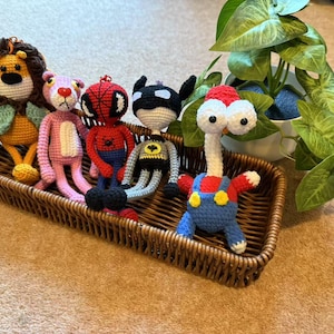 Op de afbeelding: Een verzameling handgemaakte gehaakte poppen in een rieten mand. De poppen zijn onder andere een leeuw, een roze kat, Spider-Man, Batman en een personage in een overall. De poppen zijn in verschillende kleuren, waaronder geel, rood, zwart en blauw.