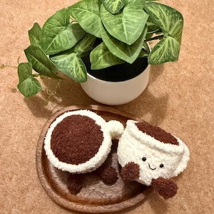 Può includere: Un set di tazza e piattino da caffè in peluche con un volto sorridente sulla tazza. La tazza e il piattino sono bianchi con dettagli marroni e poggiano su un vassoio di legno. Una pianta verde in vaso è sullo sfondo.