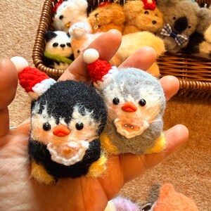 May include: Handmade felt penguin ornaments, one black and yellow, the other gray and white, both wearing tiny red Santa hats. Other felt animal ornaments, including a panda, koala, and dogs, are in a woven basket in the background.