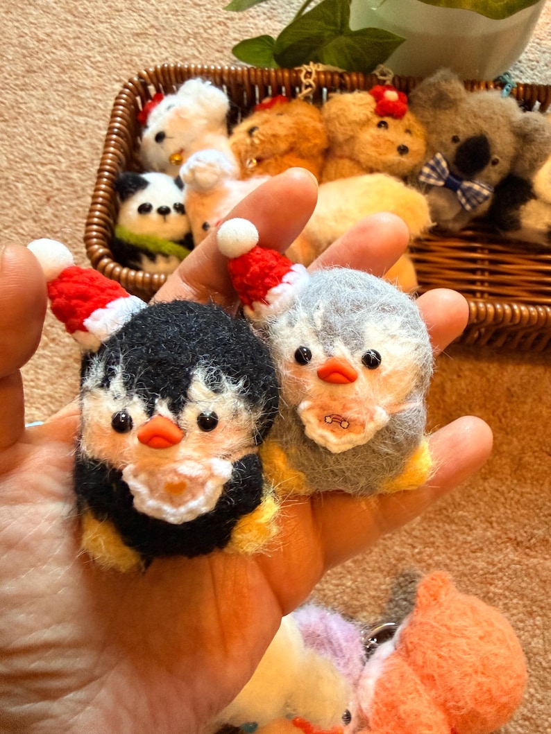 May include: Handmade felt penguin ornaments, one black and yellow, the other gray and white, both wearing tiny red Santa hats. Other felt animal ornaments, including a panda, koala, and dogs, are in a woven basket in the background.