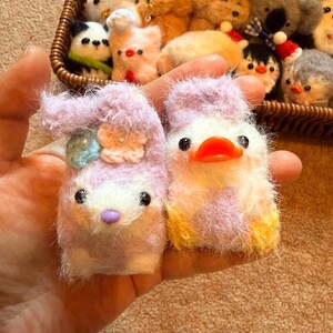 May include: A pair of handmade, fluffy animal keychains. One is a lavender bunny with a flower and a blue gem, the other a white and lavender duck with an orange beak. A basket in the background holds more plush animal keychains.