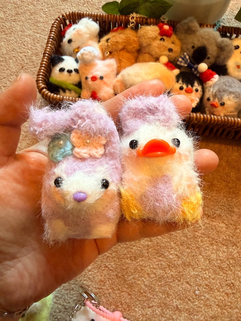 May include: A pair of handmade, fluffy animal keychains. One is a lavender bunny with a flower and a blue gem, the other a white and lavender duck with an orange beak. A basket in the background holds more plush animal keychains.