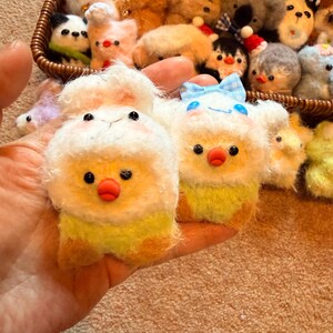 May include: Handmade felt animal keychains in a variety of designs, including chicks, pandas, and bears. The keychains are white, yellow, and brown with black eyes and red beaks. A blue bow is on one of the chicks. A wicker basket holds more keychains.