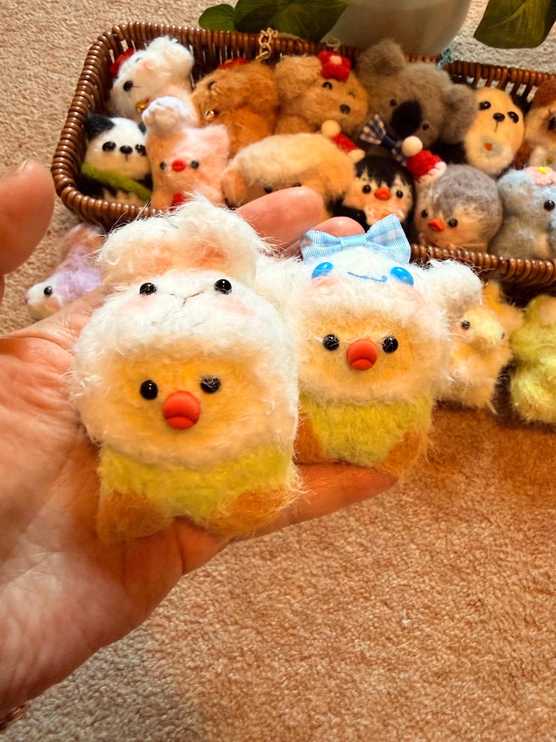 May include: Handmade felt animal keychains in a variety of designs, including chicks, pandas, and bears. The keychains are white, yellow, and brown with black eyes and red beaks. A blue bow is on one of the chicks. A wicker basket holds more keychains.