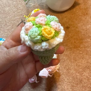 Pu&ograve; includere: Un portachiavi a bouquet all'uncinetto fatto a mano. La base a forma di cono &egrave; verde, sormontata da fiori all'uncinetto dai colori pastello. I fiori includono giallo, rosa e verde, con un bordo bianco. &Egrave; attaccato un portachiavi argentato.