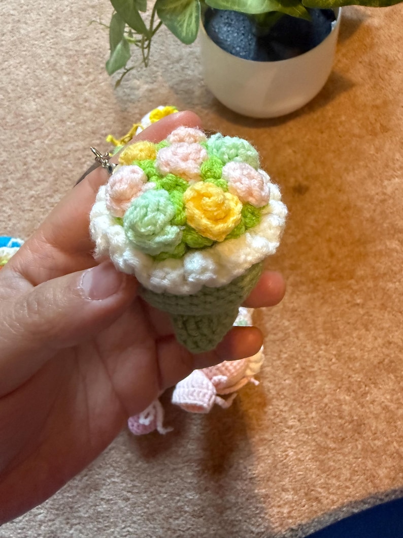 Pu&ograve; includere: Un portachiavi a bouquet all'uncinetto fatto a mano. La base a forma di cono &egrave; verde, sormontata da fiori all'uncinetto dai colori pastello. I fiori includono giallo, rosa e verde, con un bordo bianco. &Egrave; attaccato un portachiavi argentato.
