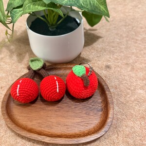 May include: Three red crocheted apple-shaped items on a wooden tray. Two are cherries, and one is a keychain. The apples have green leaves and brown stems. A small potted plant is in the background.