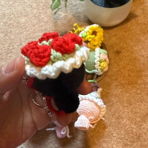 Pu&ograve; includere: Portachiavi all'uncinetto in vari design. Un portachiavi presenta una figura nera con un bouquet di fiori rossi in cima, finiture bianche e un portachiavi argentato. Altri portachiavi includono un girasole giallo, una figura verde con accenti floreali e una figura rosa.