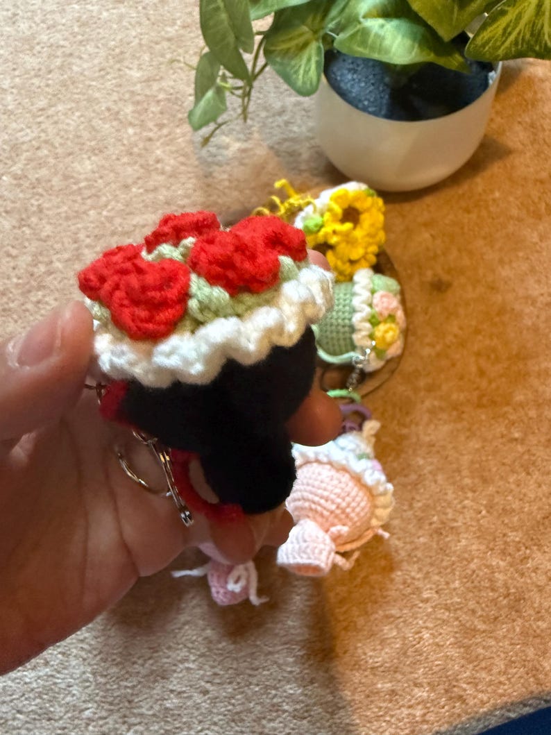Pu&ograve; includere: Portachiavi all'uncinetto in vari design. Un portachiavi presenta una figura nera con un bouquet di fiori rossi in cima, finiture bianche e un portachiavi argentato. Altri portachiavi includono un girasole giallo, una figura verde con accenti floreali e una figura rosa.