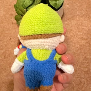 May include: A handmade crochet doll of a character wearing a green hat, blue overalls, and white gloves. The doll has a brown mustache and brown shoes. The doll is held in a hand.