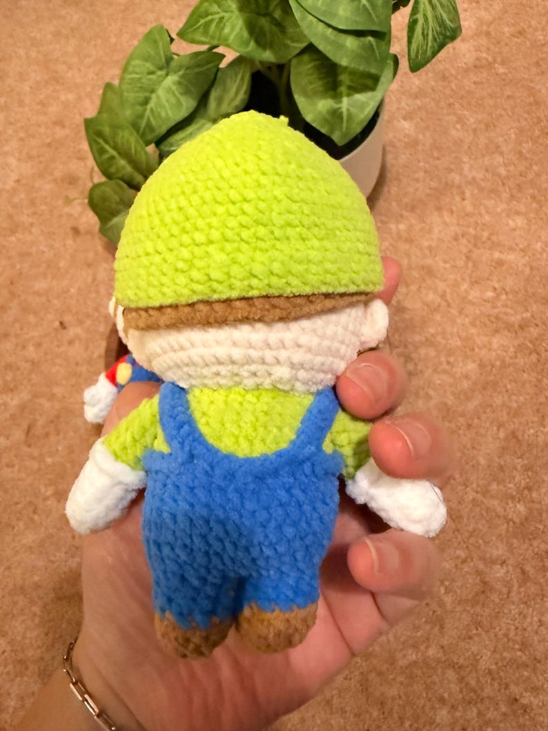 May include: A handmade crochet doll of a character wearing a green hat, blue overalls, and white gloves. The doll has a brown mustache and brown shoes. The doll is held in a hand.