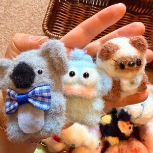 May include: A hand holds several small, handmade felt animal figures. The koala has a blue and white plaid bow tie. The duck is light blue. The dog has brown and white fur and small glasses. Other animals are visible in the background.