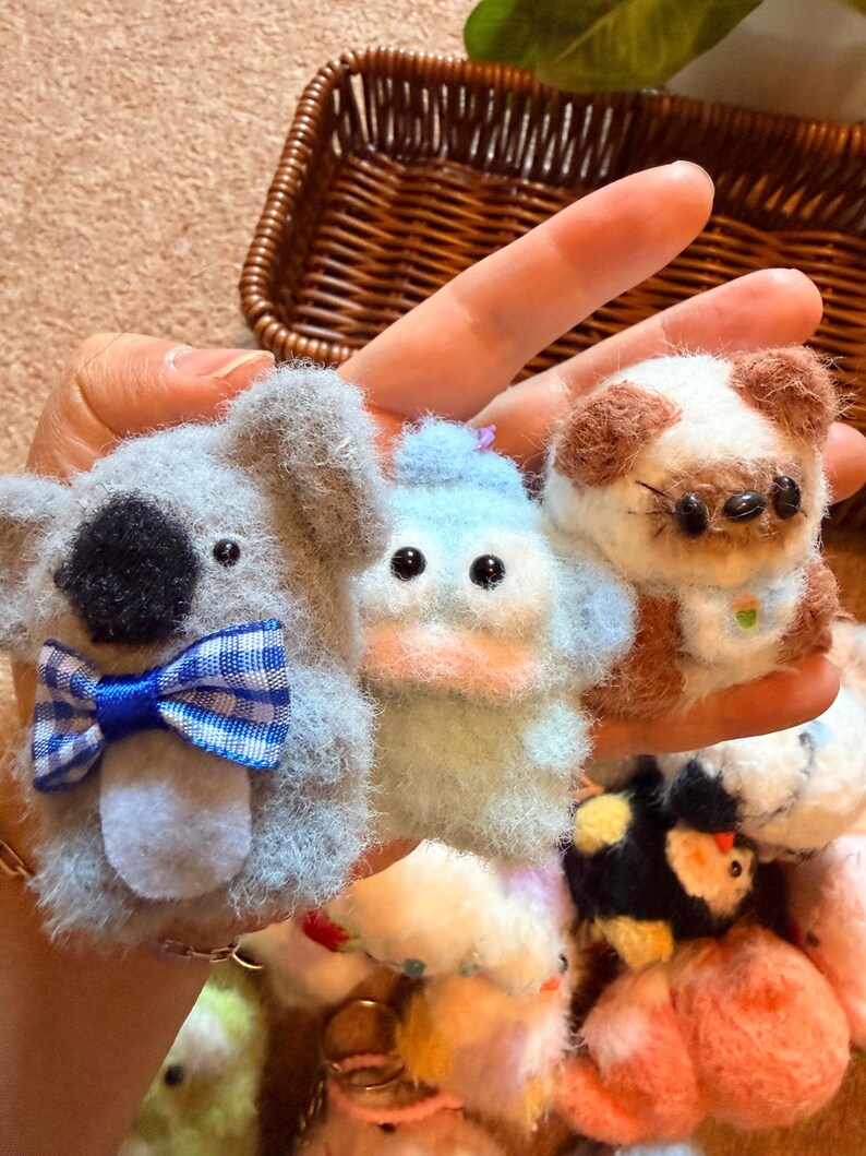 May include: A hand holds several small, handmade felt animal figures. The koala has a blue and white plaid bow tie. The duck is light blue. The dog has brown and white fur and small glasses. Other animals are visible in the background.