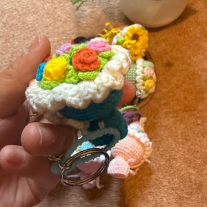 Pu&ograve; includere: Portachiavi all'uncinetto a forma di cupcake, con decorazioni floreali colorate. I cupcake hanno un bordo di glassa bianco e una base blu. I portachiavi sono attaccati ad anelli argentati.