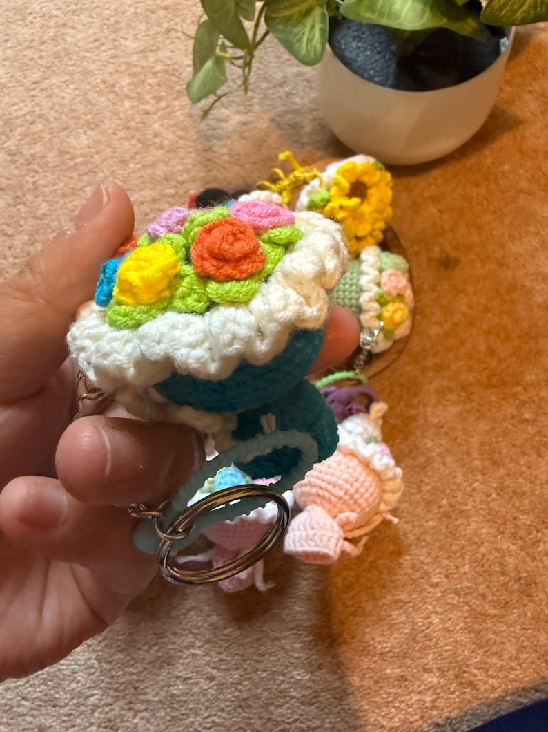 Pu&ograve; includere: Portachiavi all'uncinetto a forma di cupcake, con decorazioni floreali colorate. I cupcake hanno un bordo di glassa bianco e una base blu. I portachiavi sono attaccati ad anelli argentati.