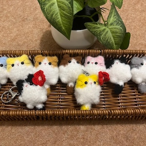 Pode incluir: Uma coleção de chaveiros artesanais em forma de gato em uma cesta de vime. Os chaveiros são brancos com vários detalhes coloridos, incluindo azul, amarelo, marrom, rosa, preto e cinza. Alguns têm enfeites de flores vermelhas. Uma planta de casa está no fundo.
