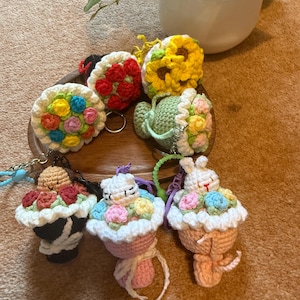 Pu&ograve; includere: Una collezione di portachiavi all'uncinetto fatti a mano in vari design. I portachiavi includono un girasole, una teiera e disegni a forma di cupcake con accenti floreali. La tavolozza dei colori comprende giallo, rosso, rosa e verde.