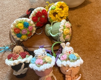 May include: A selection of handmade crochet keyrings in various designs. The keyrings include a sunflower, a teapot, and cupcake-shaped designs with floral accents. The colour palette includes yellow, red, pink, and green.