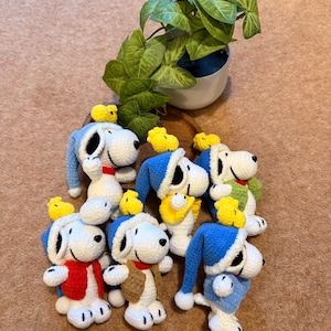 May include: A collection of handmade crochet Snoopy figures, each with a unique outfit and a small yellow bird. The Snoopy figures are primarily white with black accents, wearing blue, red, and brown clothing. A potted plant is in the background.